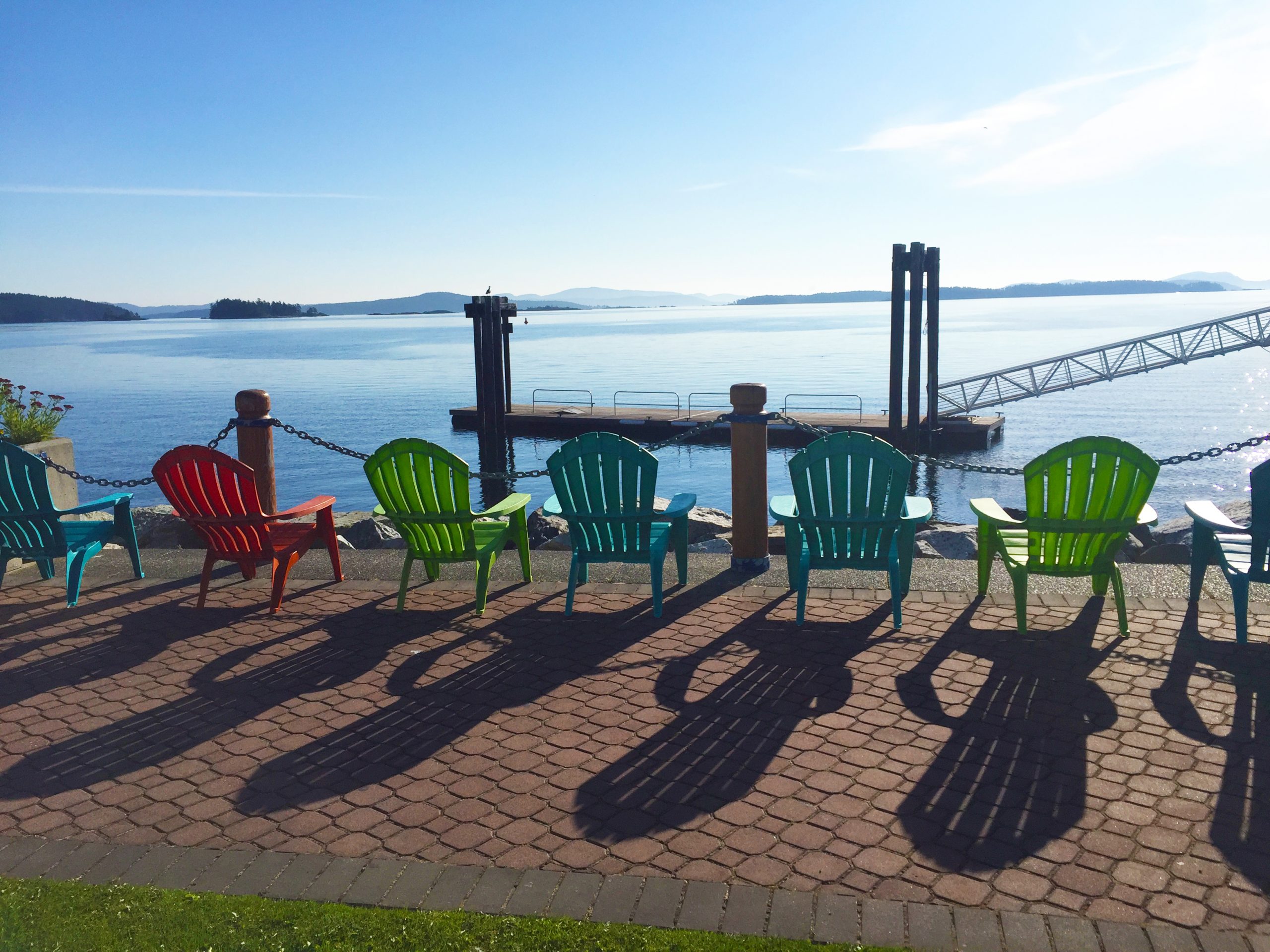 Blue, green and red Muskoka style chairs lined up along the walkway that follows the Beacon Park waterfront, with a blue sky and blue Salish Sea in the background.