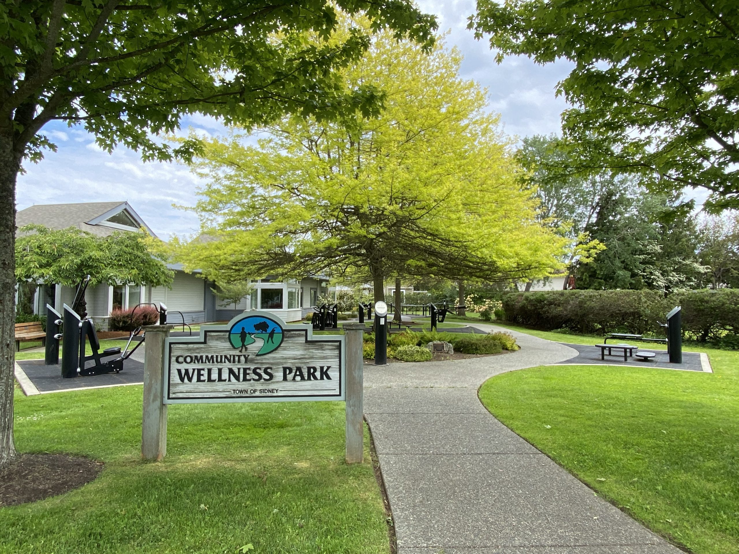 Image of Community Wellness Park with a park name sign, sidewalk, and leafy trees in the foreground and a variety of exercise equipment in the background