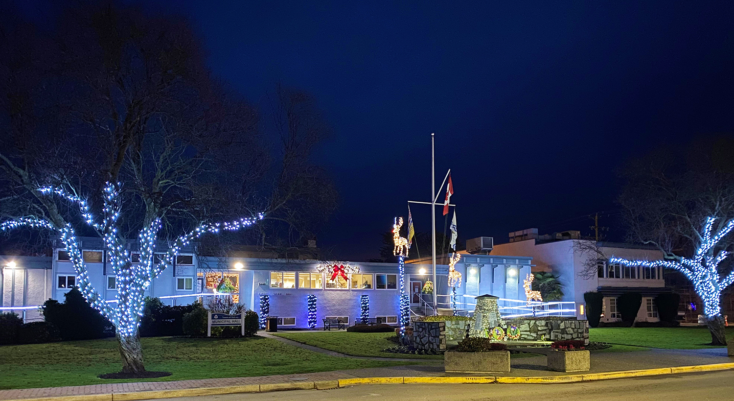 Image of the Sidney Municipal Hall building at night with Christmas lights on the trees out front and three light up reindeer on the lawn behind the cenotaph