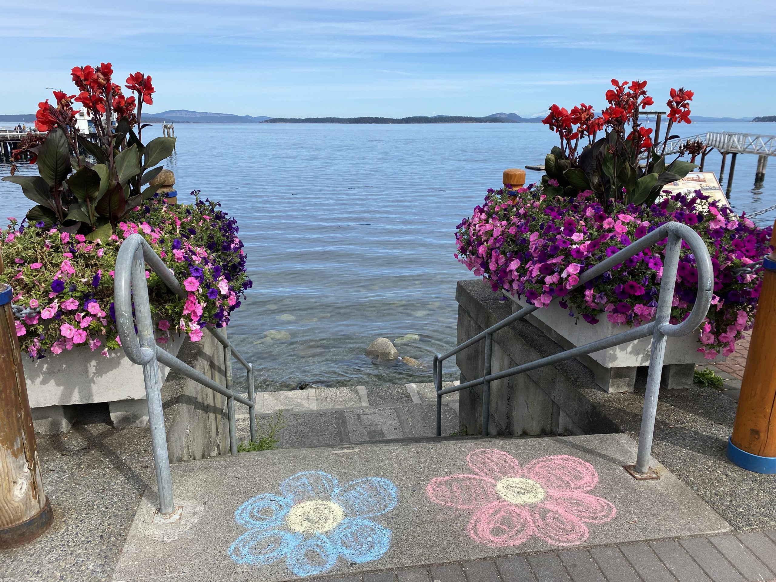 Image of the top of the stairway that leads down to Glass Beach, with planters full of pink, purple and red flowers, two flowers drawn in chalk on the sidewalk above, and blue sky and sea in the background