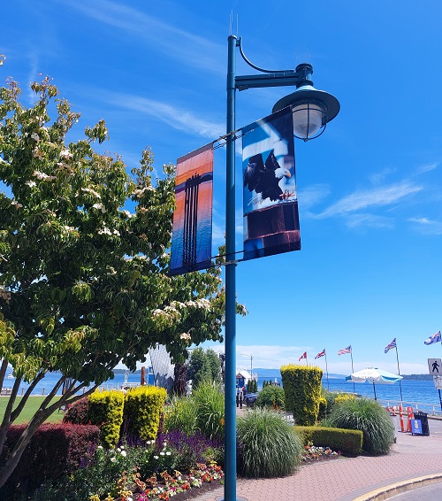 A blue lamppost with two rectangular banners on it. The left banner has a sunset image, orange and blue background of the water and islands, and the shadowed image of birds sitting on a pylon in the water. The right image is of an eagle landing on a pylon in the daytime. Behind the banners and lamppost are flowering trees and shrubbery along the sidewalk. Beacon Wharf in the background is lined with flags, and there glimpses of bright blue water, all set on a backdrop of bright blue sky.