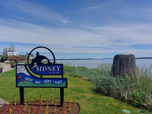 A Sidney by the sea sign in the foreground, with green space, water, and islands in the background. The sign has a breaching whale on it and says "A great lace to Shop Eat Stay Play".