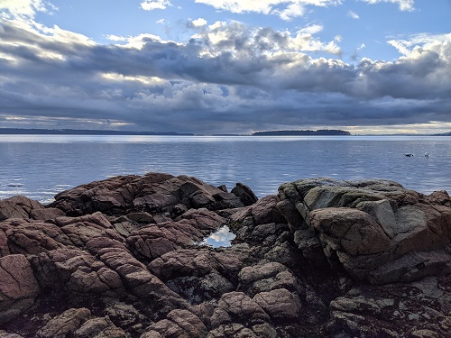 A small tide pool in the midst of large beach rocks shows a perfect reflection of the bright blue sky and moody clouds.