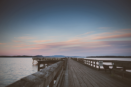 Looking down the wooden Bevan Fishing Pier towards the water at sunset, there are low fluffy clouds, reminiscent of pink and orange cotton candy, with the shadowed outlines of people walking at the end.