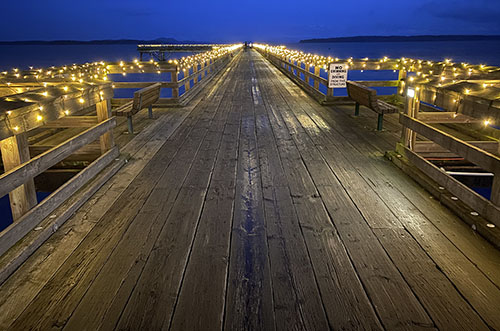 A long wooden fishing pier at night, with white twinkle lights along the railings.