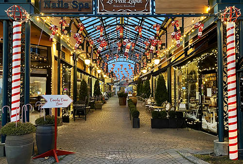 A small sign that says "Candy Cane Lane" points to a covered pedestrian street at night, which is decorated with white twinkle lights, hanging candy canes, and pillars wrapped like candy canes.