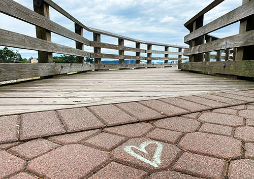 A close up of a white chalk heart on pavers, leading up to a wooden bridge.