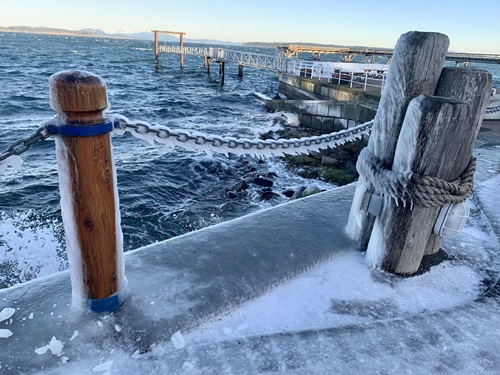 A very frosty and frozen railing of bollards and chains overlooking Glass Beach and Diver's Point. There is ice hanging off the chain, and everything facing the water is covered in a sheen of ice. The water is choppy and rough looking.