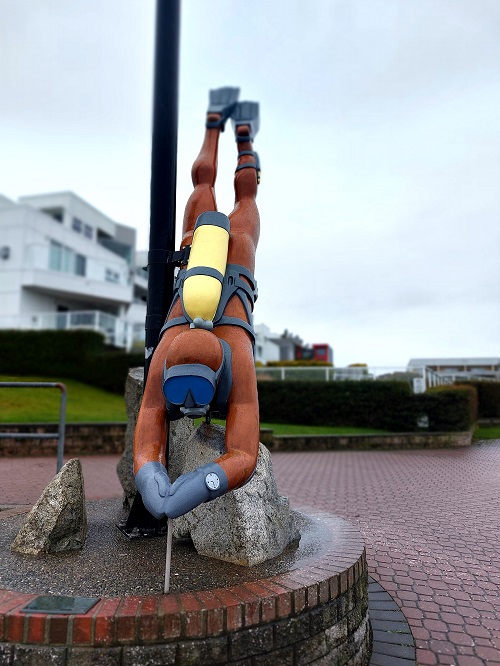 A larger-than-life 3D wooden carving of a diver, complete with air tank and mask, looks as though it is diving into in the water. It's not, though. is on a raised brick area, surrounded by a brick walking path.