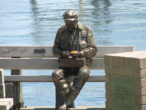 A statue of a fisherman with a tacklebox in his lap sitting on a stone bench. He is looking at his tacklebox, or perhaps it is the fresh wildflowers someone has placed in his hands.