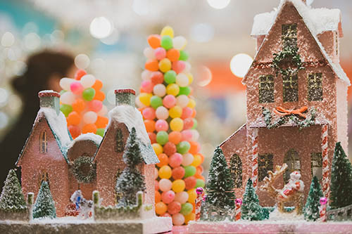 Two small decorative houses, one short and one tall, that look like they could be made of gingerbread, create a festive scene. They are covered in fake snow and have small bottle brush trees in the yards. There are two cone trees in the background, made from brightly coloured round candies.