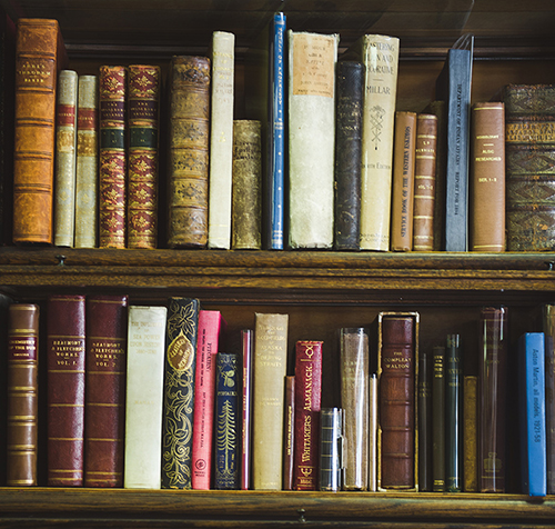 A close up photo of two shelves on a wooden book case, filled with antique looking books of various heights and colours.