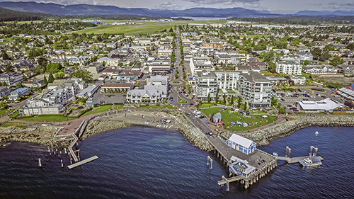 An aerial view of the Town of Sidney from over the water, facing west. The Beacon Wharf and Fishing Pier are most prominent. Beacon Avenue is centred and the airport is visible in the background.