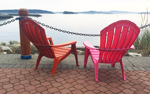 Two red Adirondack chairs sit on a cobblestone pathway, facing the waterfront. They look out over the water, with islands in the background.