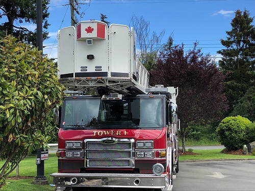 Tower 1, a shiny red ladder truck with its name in gold letters on the front, faces forward. There is a Canadian flag on the front of the white ladder.