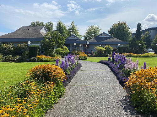 A paved path, flanked by well-tended orange and purple flowers leads up to the Sidney/North Saanich Library, a single storey blue building with a grey roof. It is a bright day, the on the other side of the flower beds is very freen, and the blue sky has wispy white clouds in it.