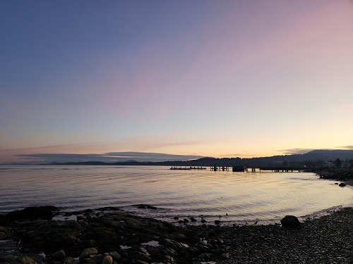 A shadowy, rocky waterfront at sunrise, with the black outline of the Anacortes Ferry Terminal. The soft orange hues of the approaching sun are visible, and the sky is various shades of blue, with wispy pink clouds.